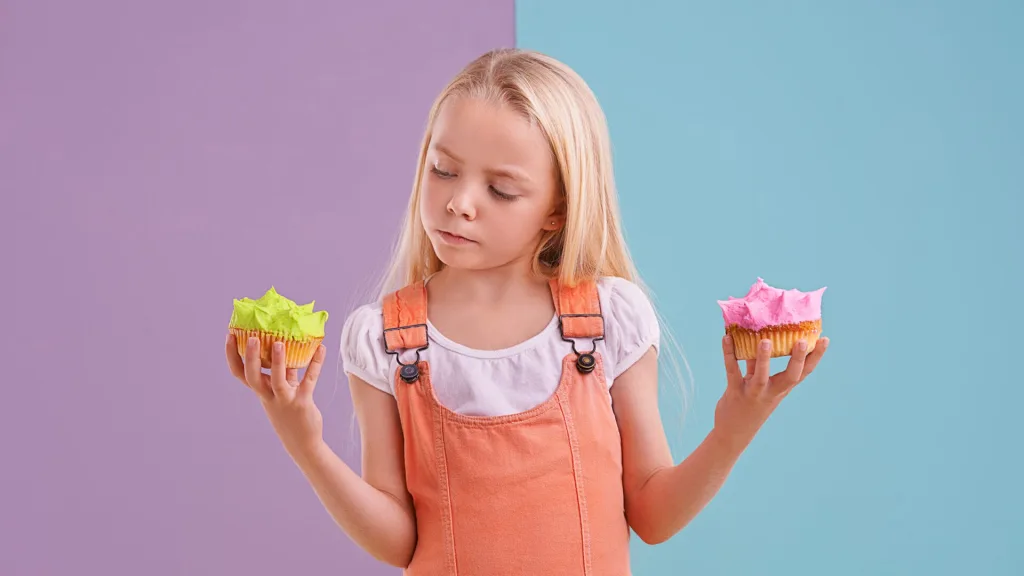緑とピンクのカップケーキを両手に持ち、どちらを食べようか悩んでいる女の子